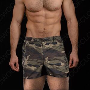 A fit guy freeballing with visible abs wearing camo print shorts stands against a dark background, with only his torso and upper legs visible.