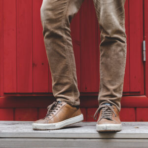 Close-up of a person's lower legs and feet wearing brown WOOF Commando Corduroy 4 inch Inseam Men's Shorts (Mesh-Lined, Button-Fly) and beige pants, freeballing, standing on a wooden surface in front of a vibrant red door.