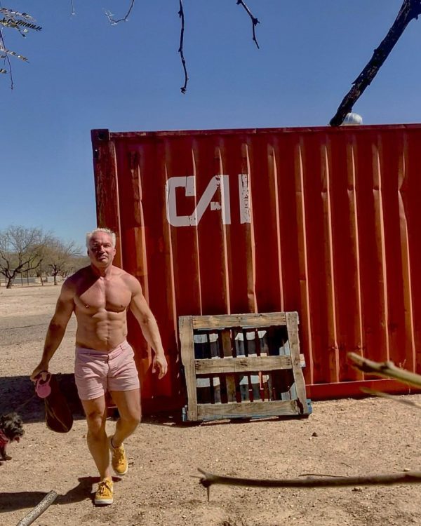 A man in WOOF Weightless Anywear™ Nylon Spandex Gym & Swim Shorts with Side Pockets and yellow shoes, choosing to go commando, carries sandals while running past a red shipping container in a dry landscape with sparse vegetation.