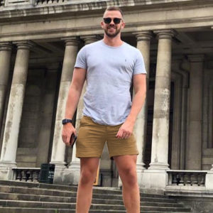 A guy in sunglasses, a gray t-shirt, and WOOF Commando Safe Chino Men's Short Shorts stands smiling in front of an ornate historical building with large columns, clearly embracing the concept of freeballing.