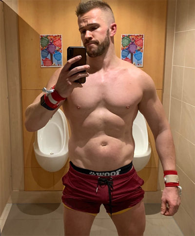 A guy in red shorts is taking a mirror selfie in a restroom, featuring urinals in the background. He has a muscular build and is wearing wristbands, holding a smartphone, and opting to free WOOF Commando Safe Jockstrap (2nd Generation Generation, Black).