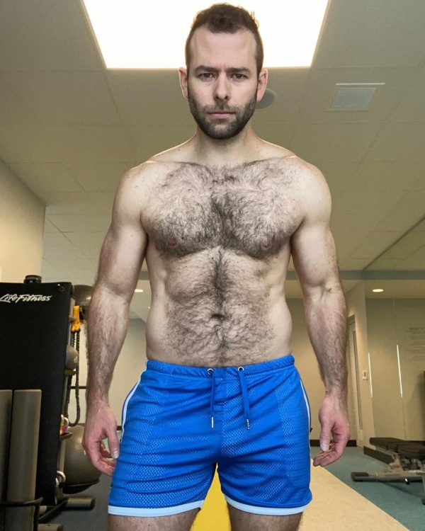 A muscular guy with a beard stands in a gym, wearing WOOF Holographic Mesh Gym Shorts (FINAL SALE!) and looking intently at the camera. His physique is well-defined, highlighted by natural lighting from above, with noticeable bulge enhancement.