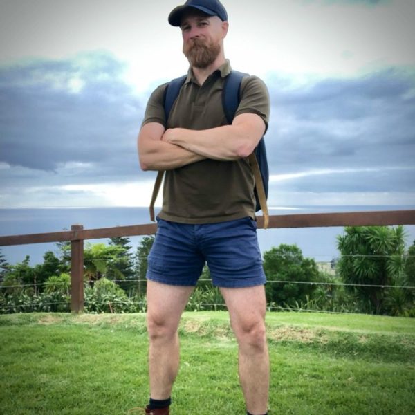 A guy with a beard stands confidently with his arms crossed in a park, wearing a green shirt, WOOF Original Mesh-Lined 4" Inseam Chinos, denim shorts while freeballing, and a backpack under an overcast sky.
