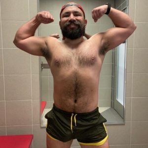 A bearded guy freeballing and flexing his muscles in a gym locker room, wearing WOOF Holographic Mesh Gym Shorts (FINAL SALE!), with a humorous expression.