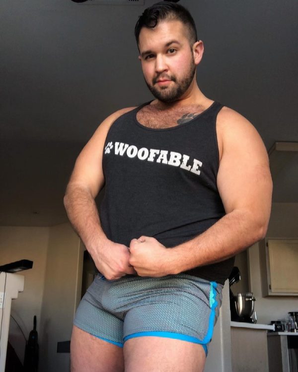 A guy with a beard stands indoors, smiling as he holds the edge of his WOOF Holographic Mesh Gym Shorts, showcasing bulge enhancement in his gray and blue outfit in a casual, home environment.