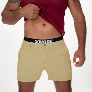 A guy lifts up his red mesh tank top to reveal his fit abdomen and a pair of light brown shorts designed for bulge enhancement, featuring a black band.