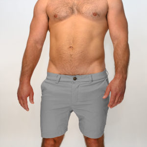 A guy stands facing the camera, wearing WOOF Commando Safe Chino Men's Short Shorts, 6 inch Inseam, Mesh-Lined and no shirt, showcasing his torso and bulge enhancement against a white background.