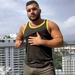 A guy in a WOOF Weightless Mesh Nylon-Spandex Men's Tank Top standing on a balcony with a cityscape in the background, go Commando, making a gesture with one hand on his chest.